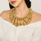 A close-up of a model wearing a chunky, wide gold statement necklace with textured, layered scales. The model is wearing an off-the-shoulder white top against a clean, light beige background.