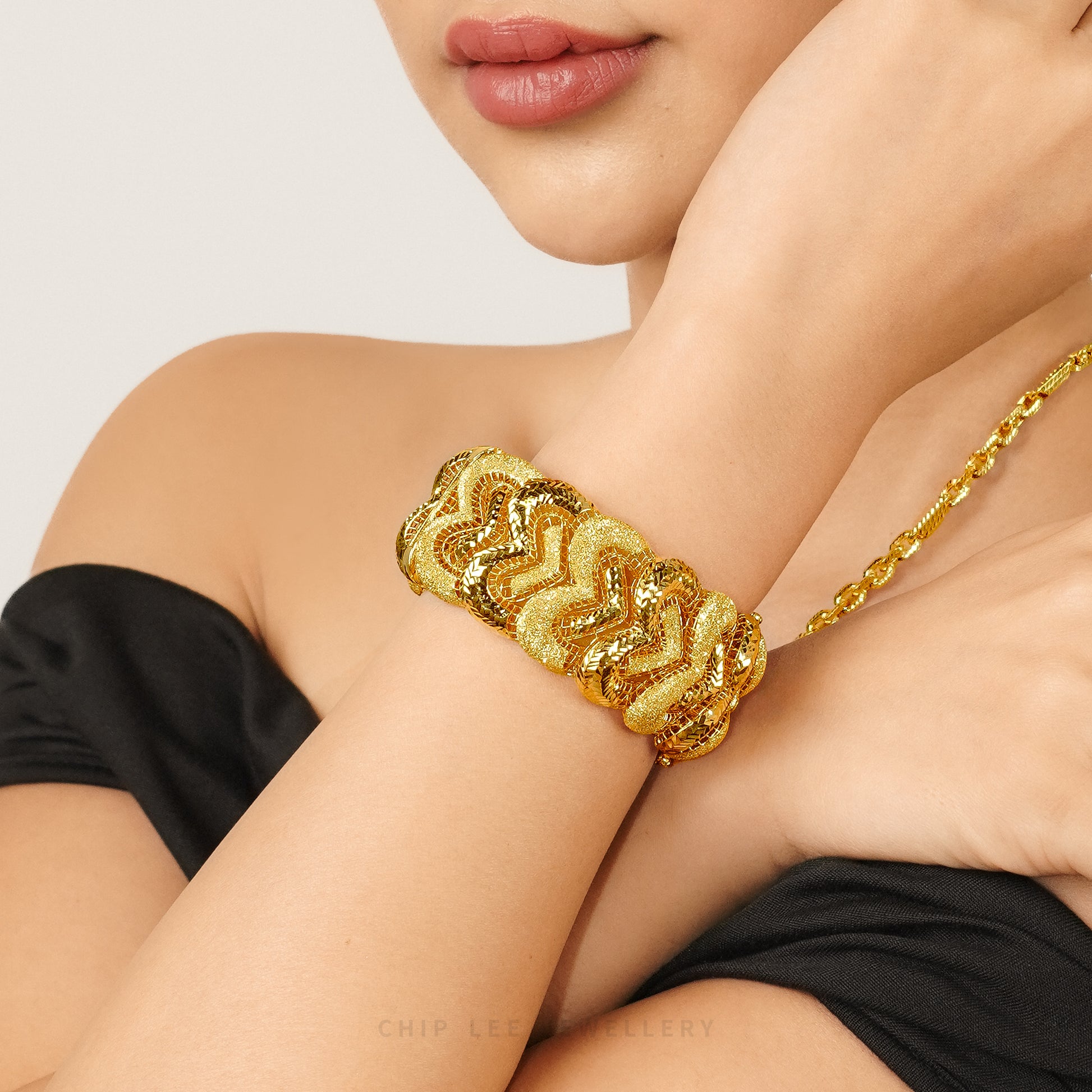 Close-up of a model wearing a wide gold bracelet with interwoven wave motifs and contrasting textured and polished finishes, styled with a black off-shoulder top.