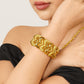 Close-up of a model wearing a wide gold bracelet with interwoven wave motifs and contrasting textured and polished finishes, styled with a black off-shoulder top.