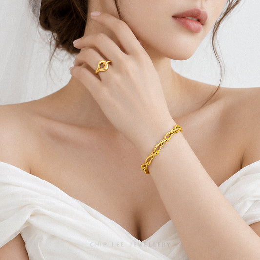 A close-up lifestyle shot of a model wearing a gold heart-shaped ring on one finger and a matching twisted gold chain bracelet on the wrist, wearing a white off-the-shoulder top.