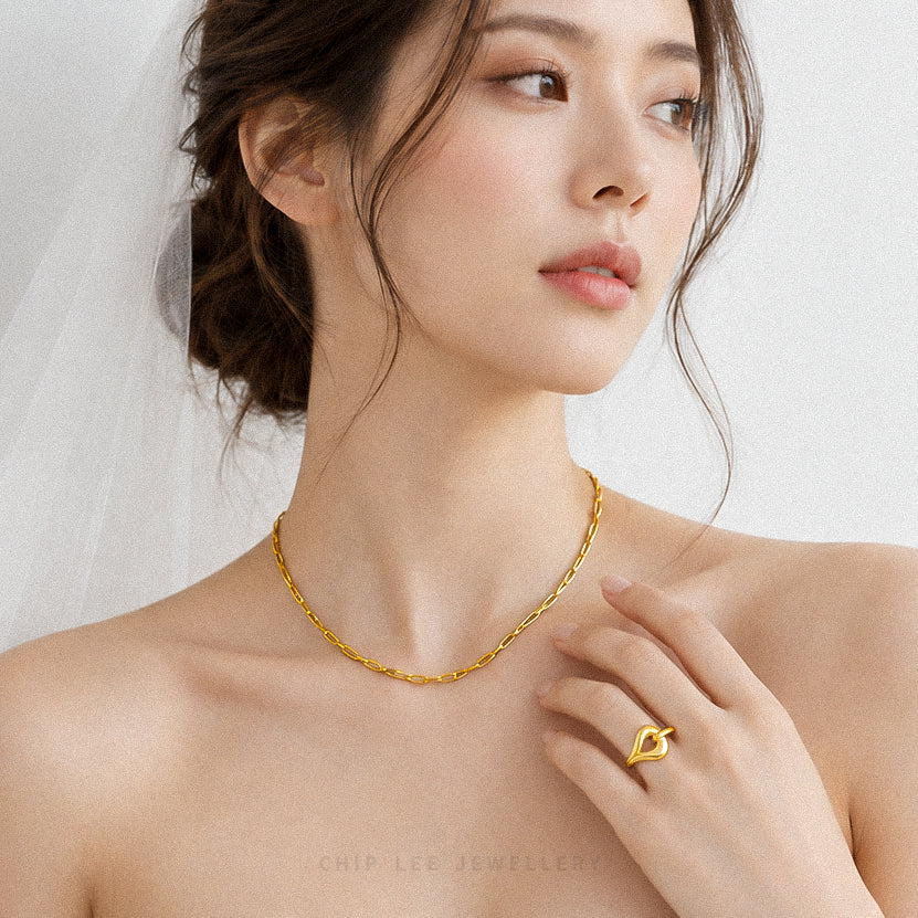A woman wearing a delicate gold chain necklace and a matching gold heart-shaped ring, shown in a soft, bridal-style close-up. The brand name 'CHIP LEE JEWELLERS' is faintly visible at the bottom.