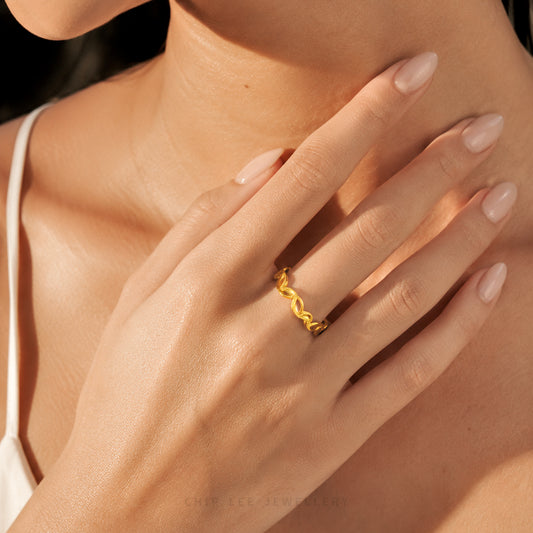 A close-up lifestyle shot of a woman's hand wearing a thin, wavy gold ring, with her hand resting on her neck. She is wearing a white spaghetti strap top, and the lighting is warm and golden.
