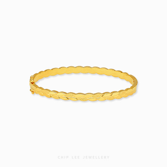 A polished yellow gold bangle bracelet with a textured, ridged outer surface and a smooth inner band, displayed on a clean white background with a brand watermark at the bottom.