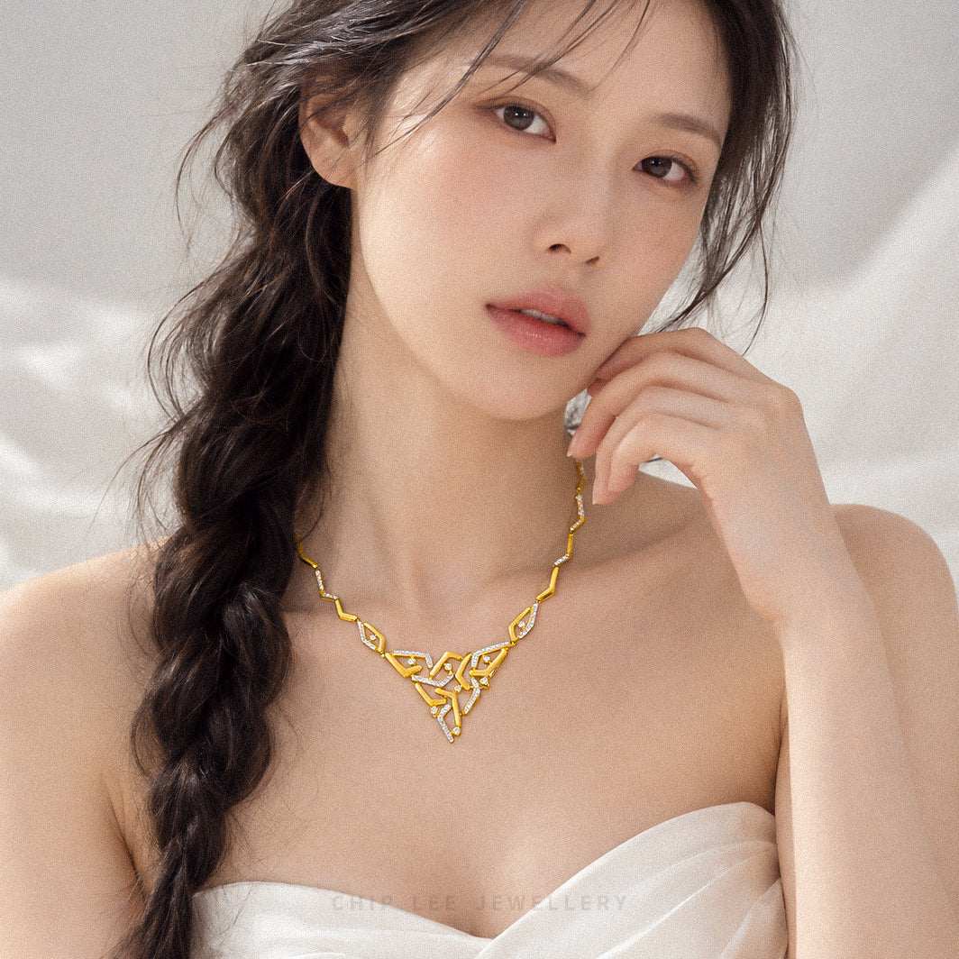 A woman with long dark hair in a side braid wears a gold necklace with an intricate openwork knot design. She is wearing a white strapless top against a soft, light gray background.