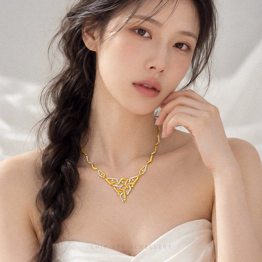A woman with long dark hair in a side braid wears a gold necklace with an intricate openwork knot design. She is wearing a white strapless top against a soft, light gray background.