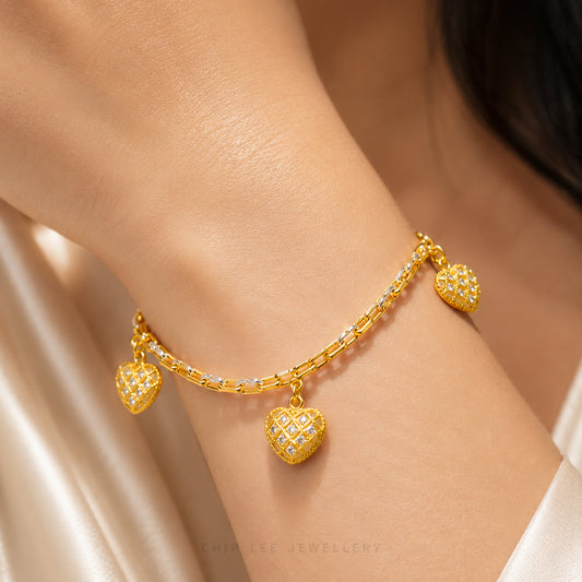 A close-up of a gold chain bracelet with three heart-shaped charms, worn on a person's wrist. The heart charms are detailed with small clear stones.