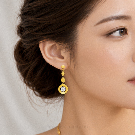 A close-up of a woman wearing a gold drop earring with a circular yellow-gold pendant and a clear central stone. The background is a soft, light neutral color.