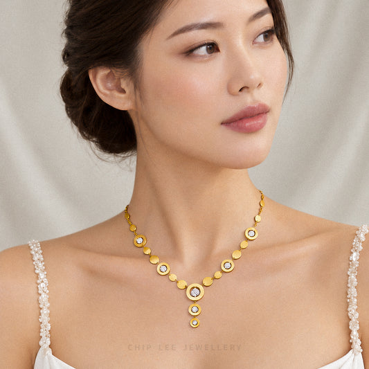 A woman wearing a gold necklace with circular disc motifs and clear rhinestone accents, photographed against a soft light beige fabric backdrop. The brand name 'CHIP LEE JEWELLERY' is faintly visible at the bottom.