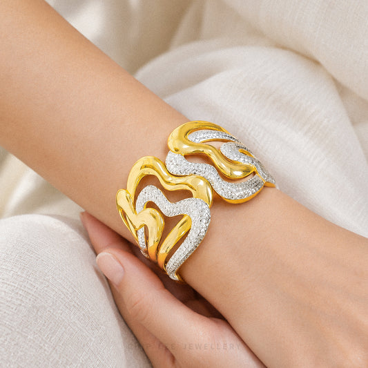 A close-up of a gold and silver wavy cuff bracelet worn on a person's wrist, set against a soft cream-colored fabric background.