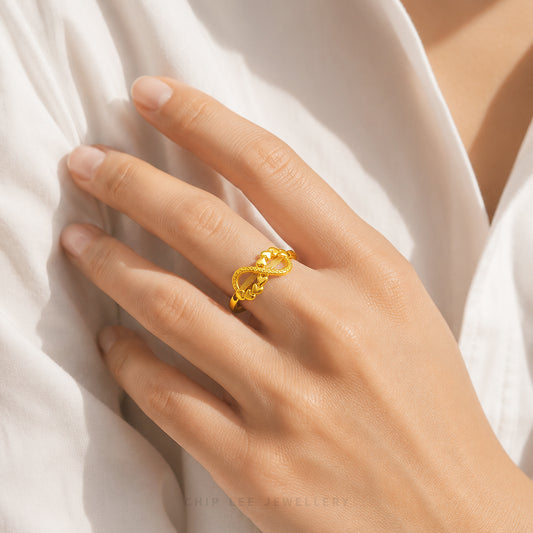 A close-up of a hand wearing a gold ring with a textured, rope-like knot design, resting against white fabric.