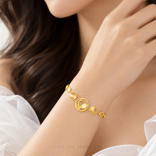 A close-up lifestyle shot of a woman's wrist wearing a delicate gold bracelet with interlocking circular and oval motifs. The background is softly blurred, showing her hand and hair, with a white sheer top visible at the edges.