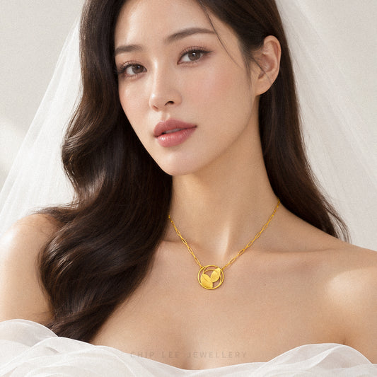 A close-up of a woman wearing a delicate gold necklace with a circular pendant featuring a stylized bird design. She is dressed in white bridal attire with a veil, and the background is a soft, neutral, light beige tone.