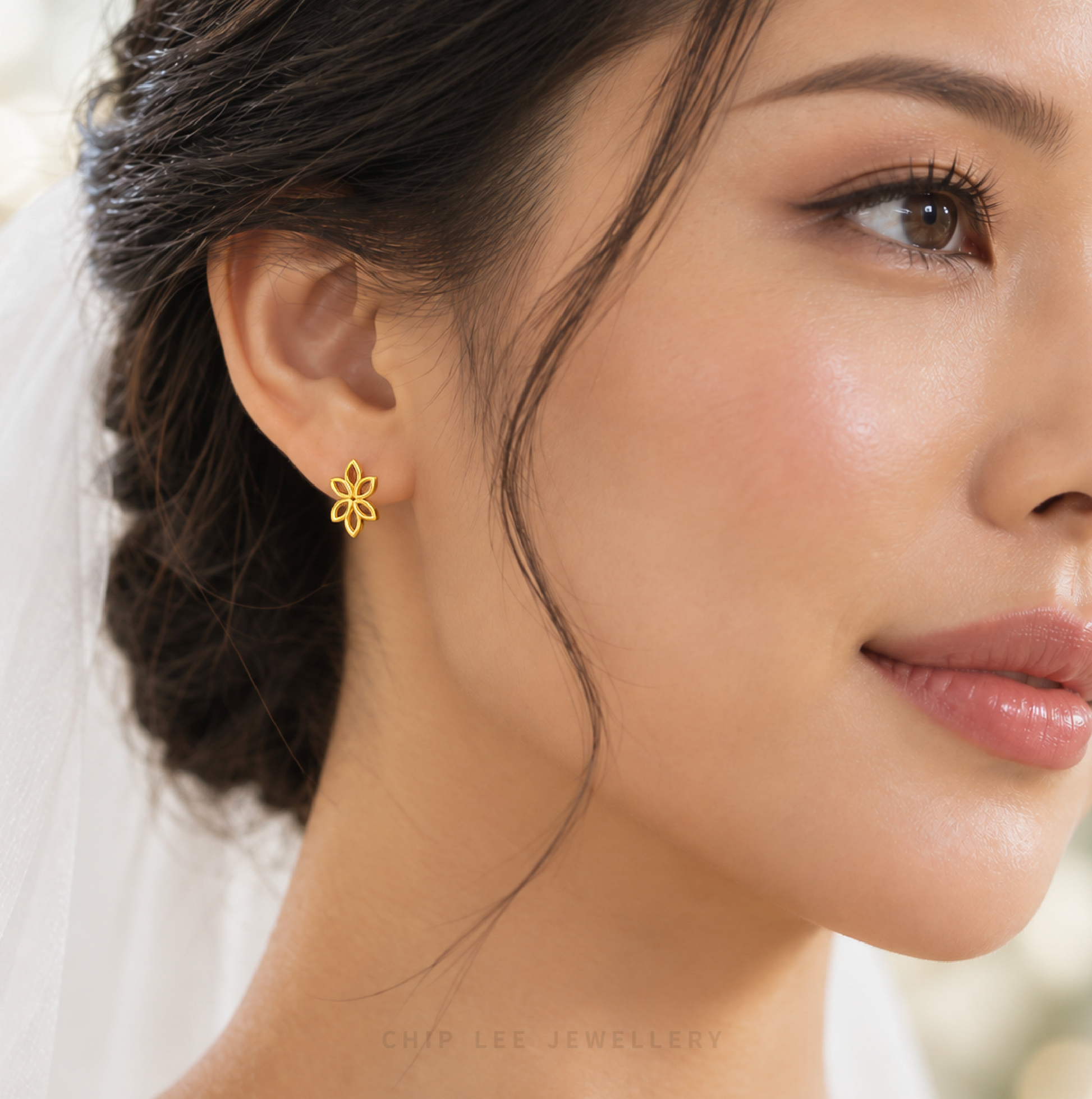 A close-up of a woman's ear wearing a delicate gold flower-shaped stud earring. The image features soft, warm lighting and a clean, blurred background with a hint of a white veil.