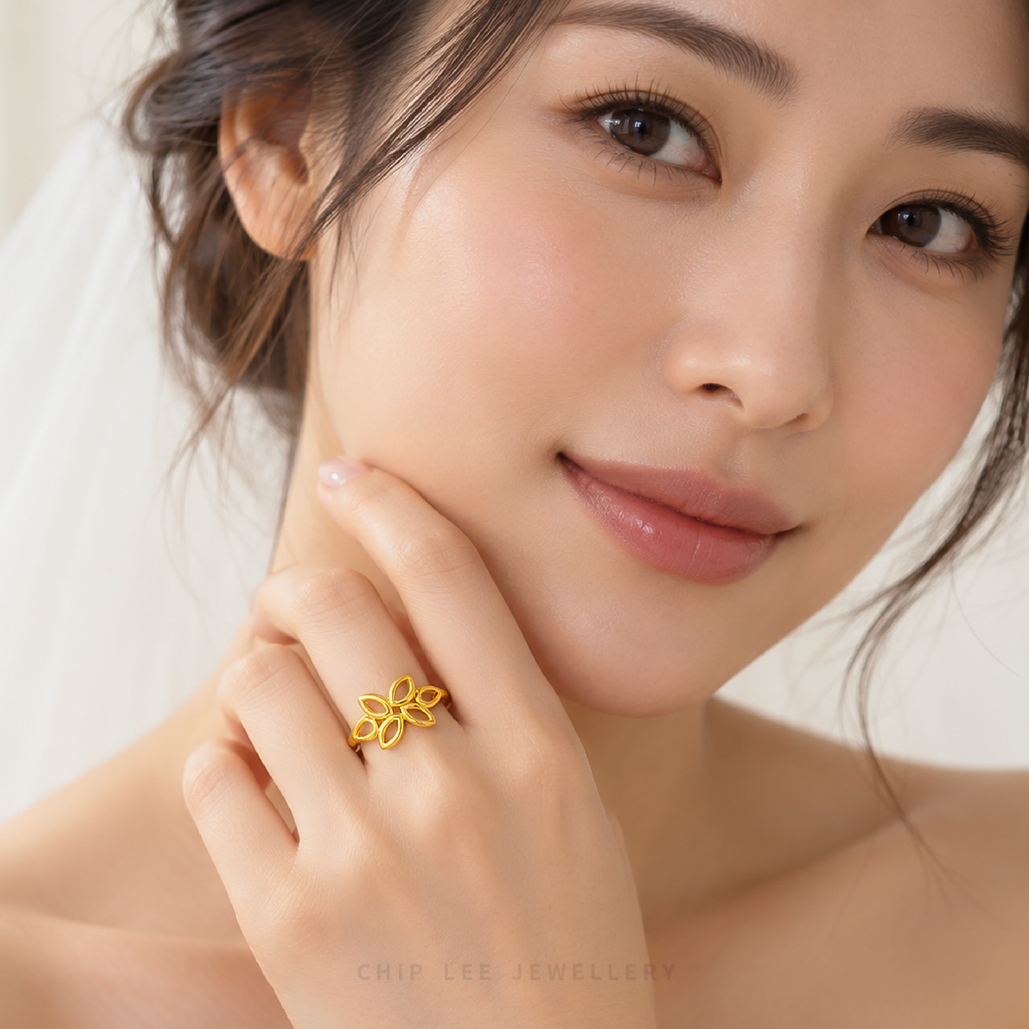 A close-up lifestyle shot of a woman wearing a delicate gold ring with a floral or butterfly-like openwork design on her finger. She is smiling gently, with soft, warm lighting and a clean, light background.