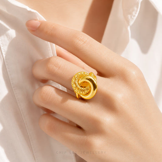 A close-up of a hand wearing a chunky, polished gold ring with a textured knot-like design, styled with a white collared shirt.