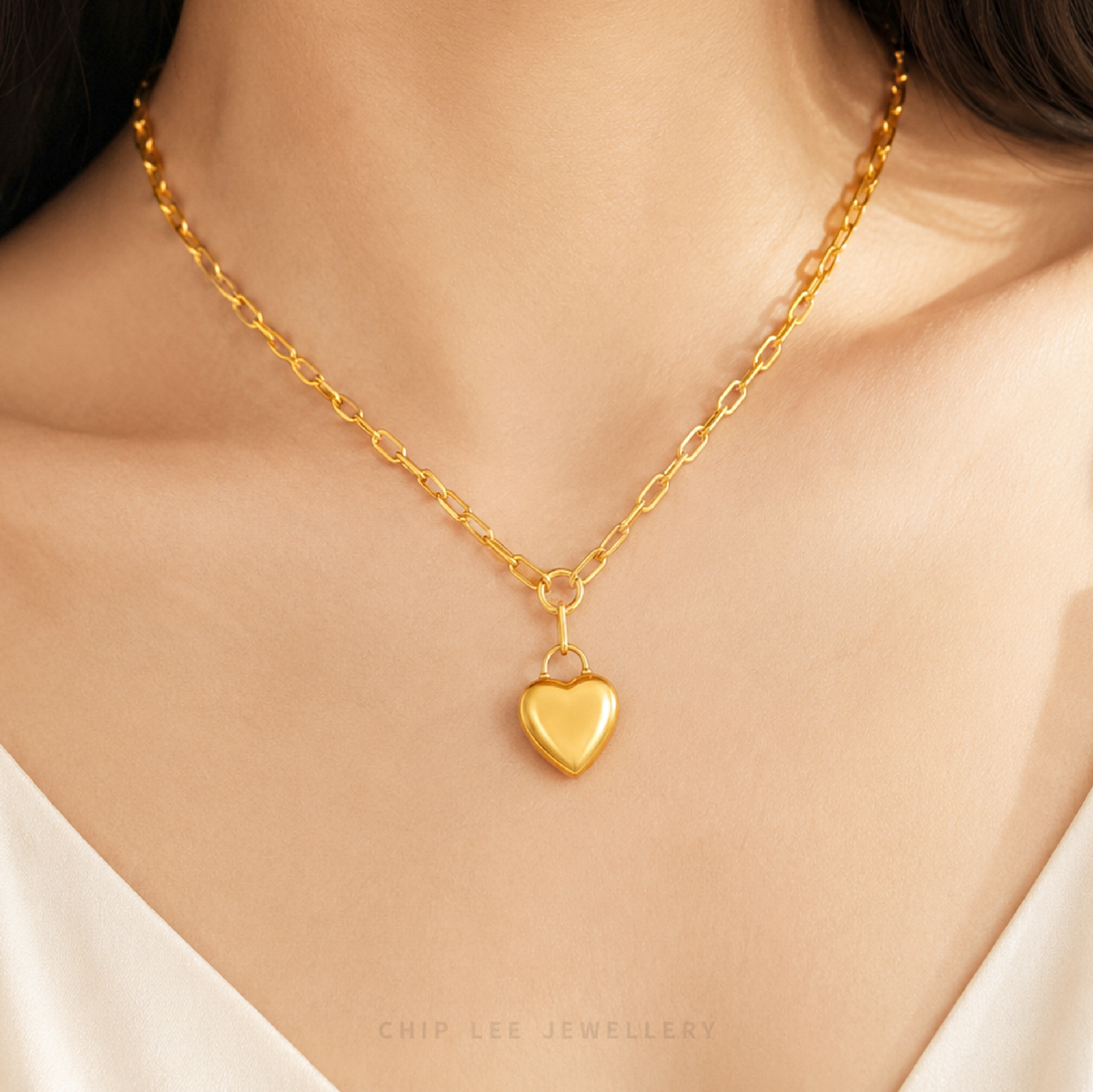 A close-up of a model's neck and chest wearing a delicate gold chain necklace with a small polished heart-shaped pendant. The image features a warm, soft glow and has 'CHIP LEE JEWELLERY' text printed at the bottom.