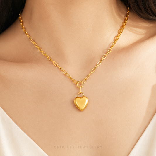 A close-up of a model's neck and chest wearing a delicate gold chain necklace with a small polished heart-shaped pendant. The image features a warm, soft glow and has 'CHIP LEE JEWELLERY' text printed at the bottom.