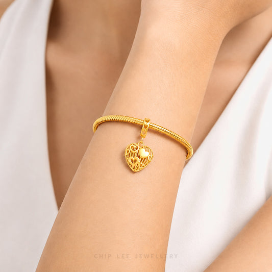 A close-up of a gold snake-bangle bracelet with a dangling filigree heart charm, worn on a person's wrist against a white garment background.