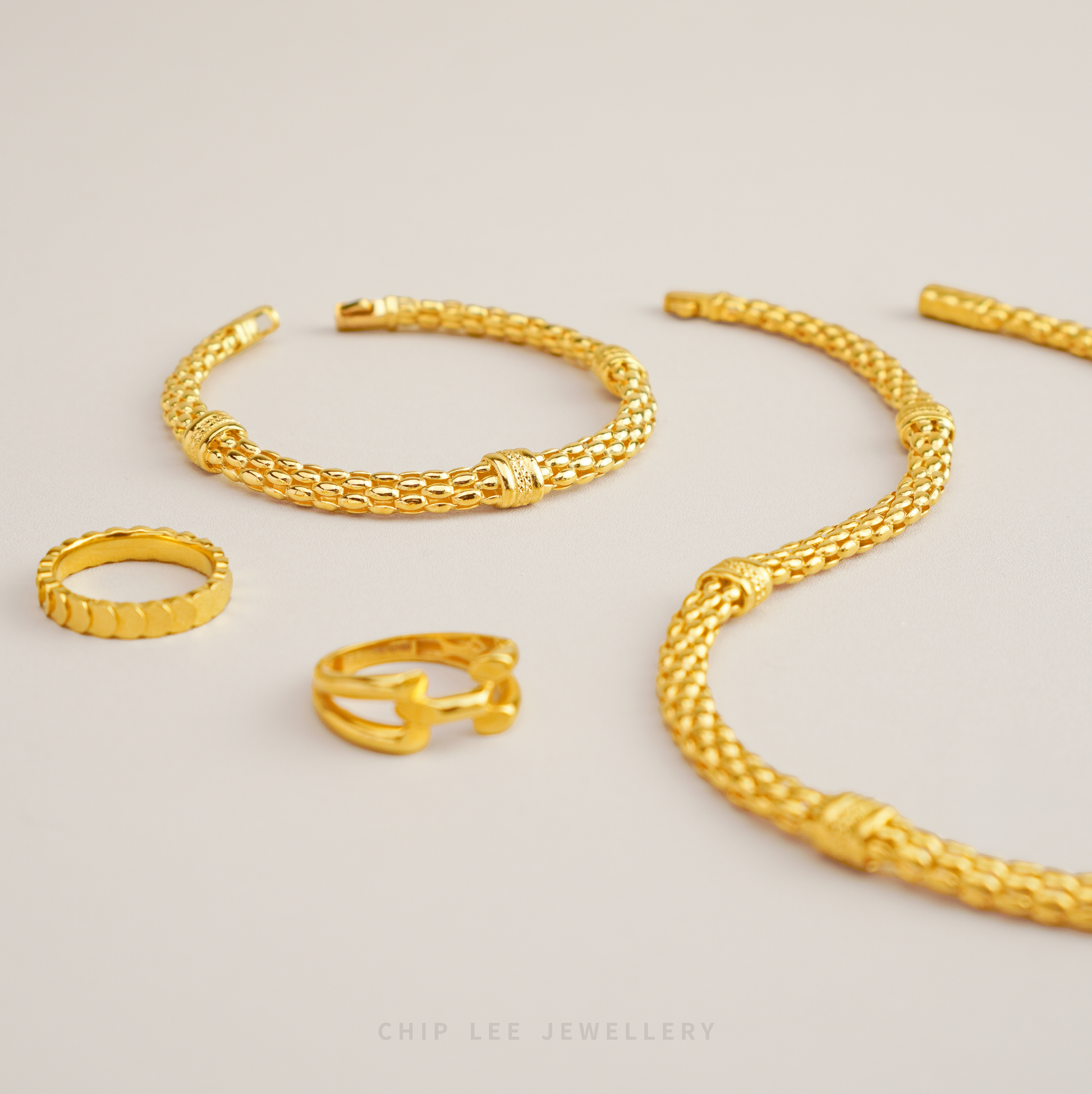 Gold jewelry including a necklace, bracelet, and rings on a light gray background.