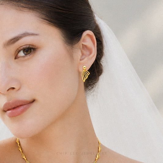 A close-up of a woman wearing a delicate gold leaf-shaped stud earring and a matching gold chain necklace, with a white bridal veil in the background.