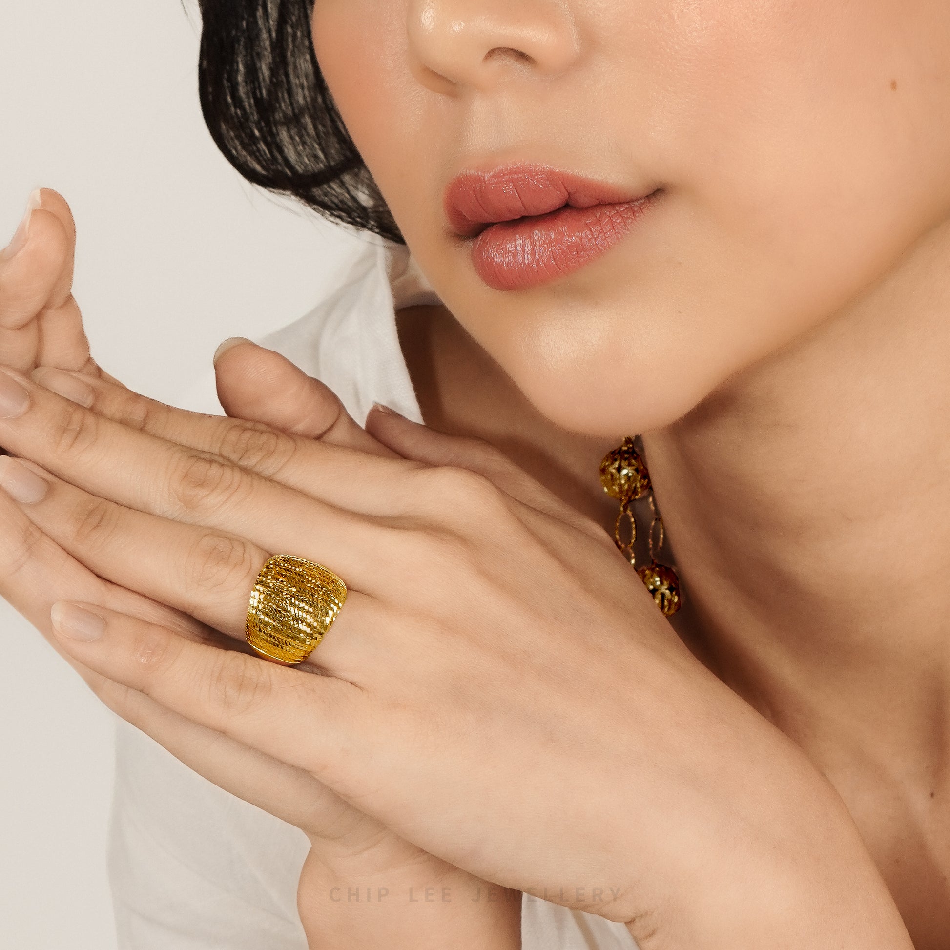 A close-up lifestyle shot of a model wearing a chunky, textured gold ring on her finger, with her hands crossed in front of her face against a clean, light background.