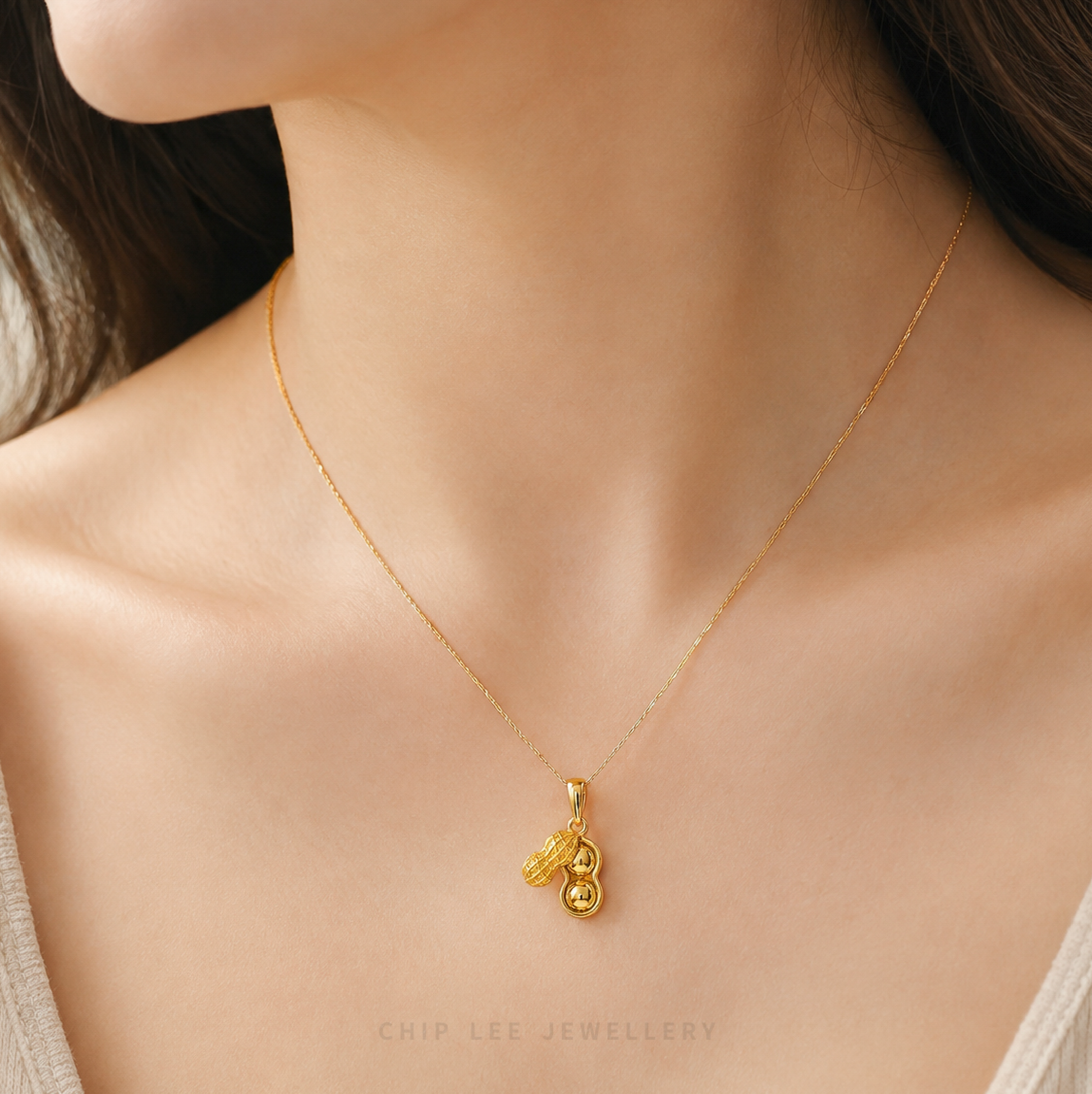 A close-up of a model's neck and chest wearing a delicate gold chain necklace with a small pendant. The pendant features a stylized gold figure with a small dangling charm.