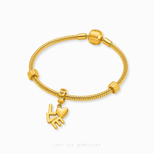 A gold-plated braided cord bracelet with a heart and hand charm, displayed on a clean white background with a brand watermark at the bottom.