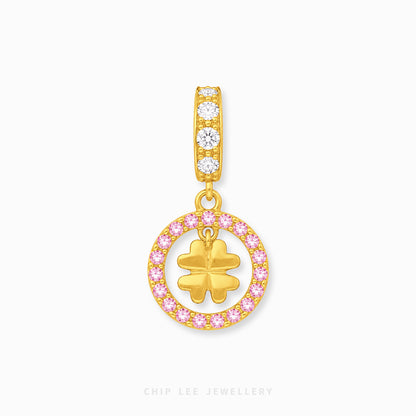 A gold charm pendant featuring a vertical bar with three clear stones and a dangling circular charm outlined in pink stones, with a gold four-leaf clover in the center, shown on a light gray background.