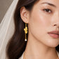 A close-up of a woman wearing a gold dangle earring with a floral motif and a small white pearl drop, paired with a white bridal veil. The brand name 'LEE JEWELLERY' is subtly visible at the bottom.