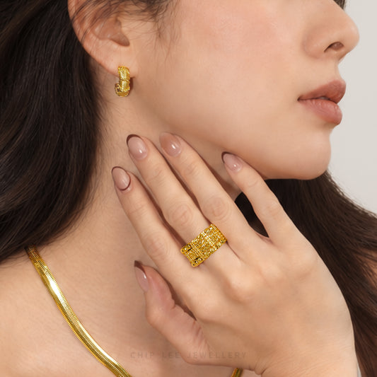 A close-up lifestyle shot of a model wearing a chunky textured gold ring on her finger, a matching gold huggie hoop earring, and a gold snake chain necklace against a light gray background.