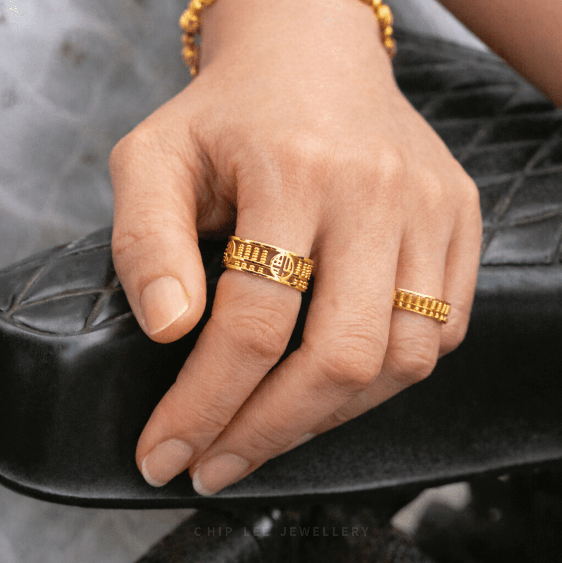 Close-up of a hand resting on a black chair, wearing 916 gold rings with intricate geometric and symbolic engravings.