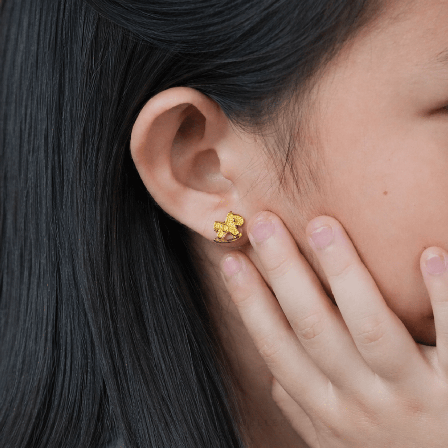 Close-up of an ear wearing a delicate 916 gold rocking horse stud earring, gently held by fingers