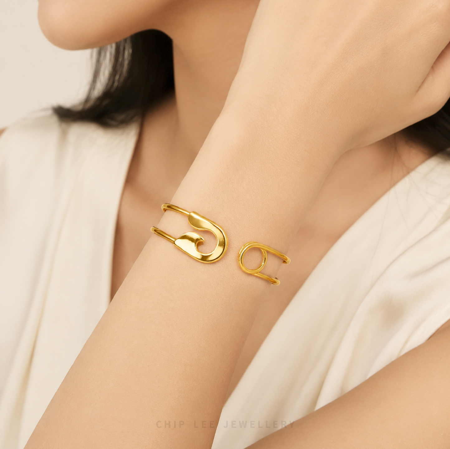 A close-up lifestyle shot of a model's wrist wearing a polished gold cuff bracelet with a safety-pin-inspired open design. The model is wearing a white satin top, and the background is a soft, neutral studio setting.
