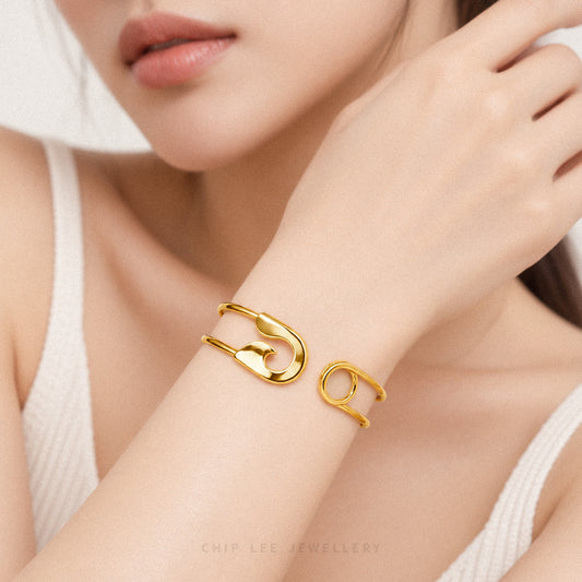 A close-up lifestyle shot of a model wearing a polished gold cuff bracelet in the shape of a safety pin. The model is wearing a white ribbed tank top, and the background is a soft, out-of-focus neutral tone.