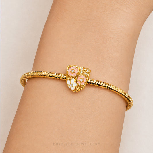 A gold snake-braided bracelet with a central shield-shaped charm featuring pink and white enamel flowers, shown worn on a person's wrist.