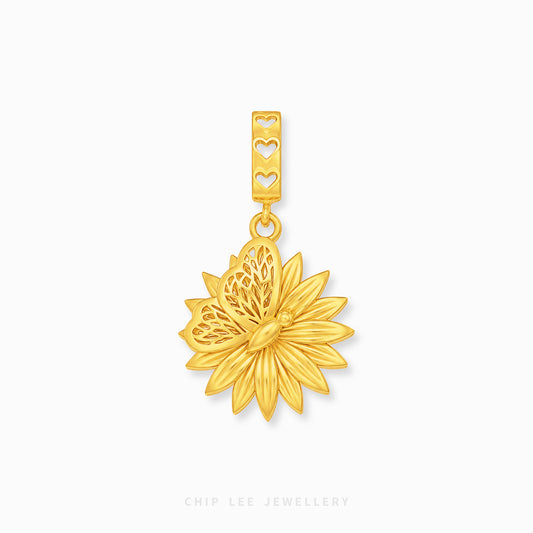 A gold flower-shaped pendant charm with a textured center and a bail featuring three heart-shaped cutouts, shown on a clean white background with a brand watermark at the bottom.