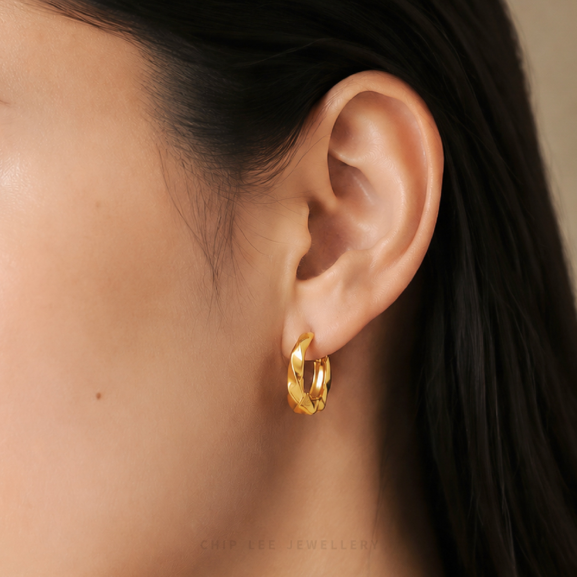 A close-up of a model's ear wearing a textured gold huggie hoop earring. The image features warm, soft lighting and a neutral beige background.