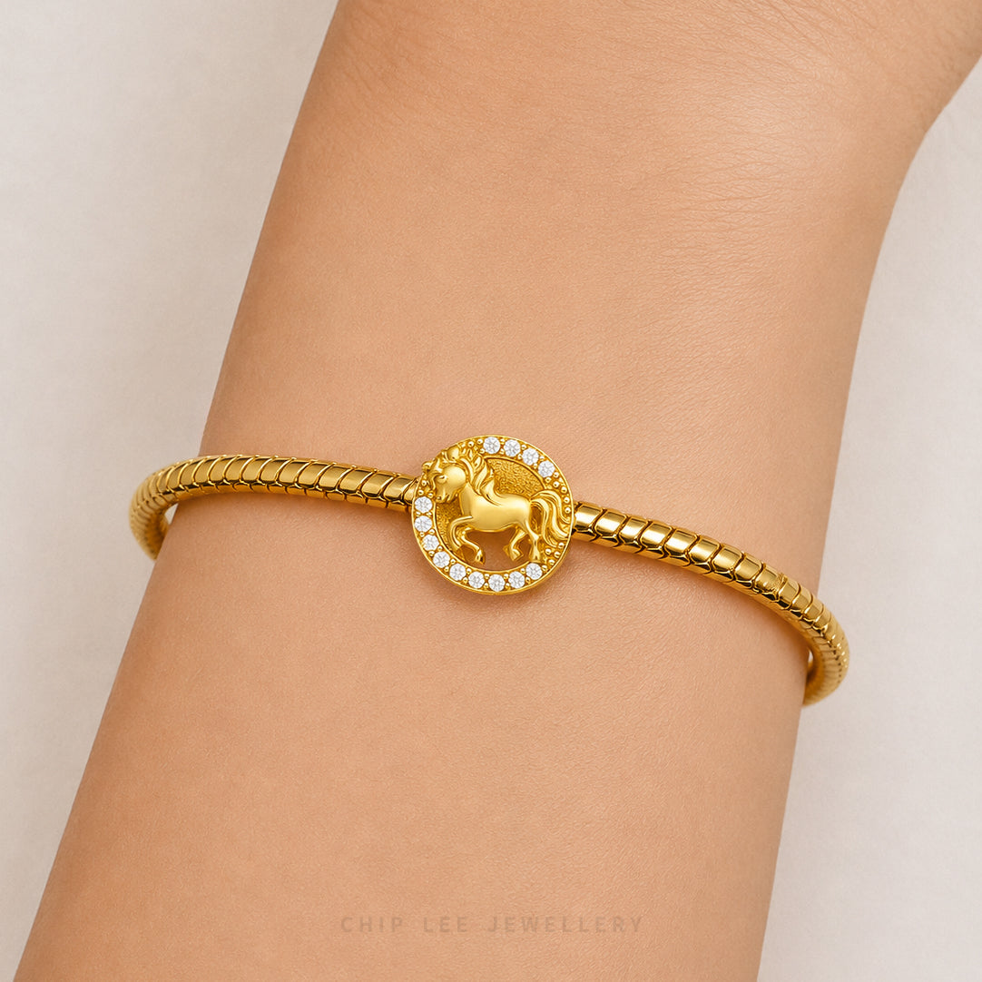 A gold-plated bangle bracelet with a central circular charm featuring a unicorn motif surrounded by small clear stones, shown worn on a person's wrist.