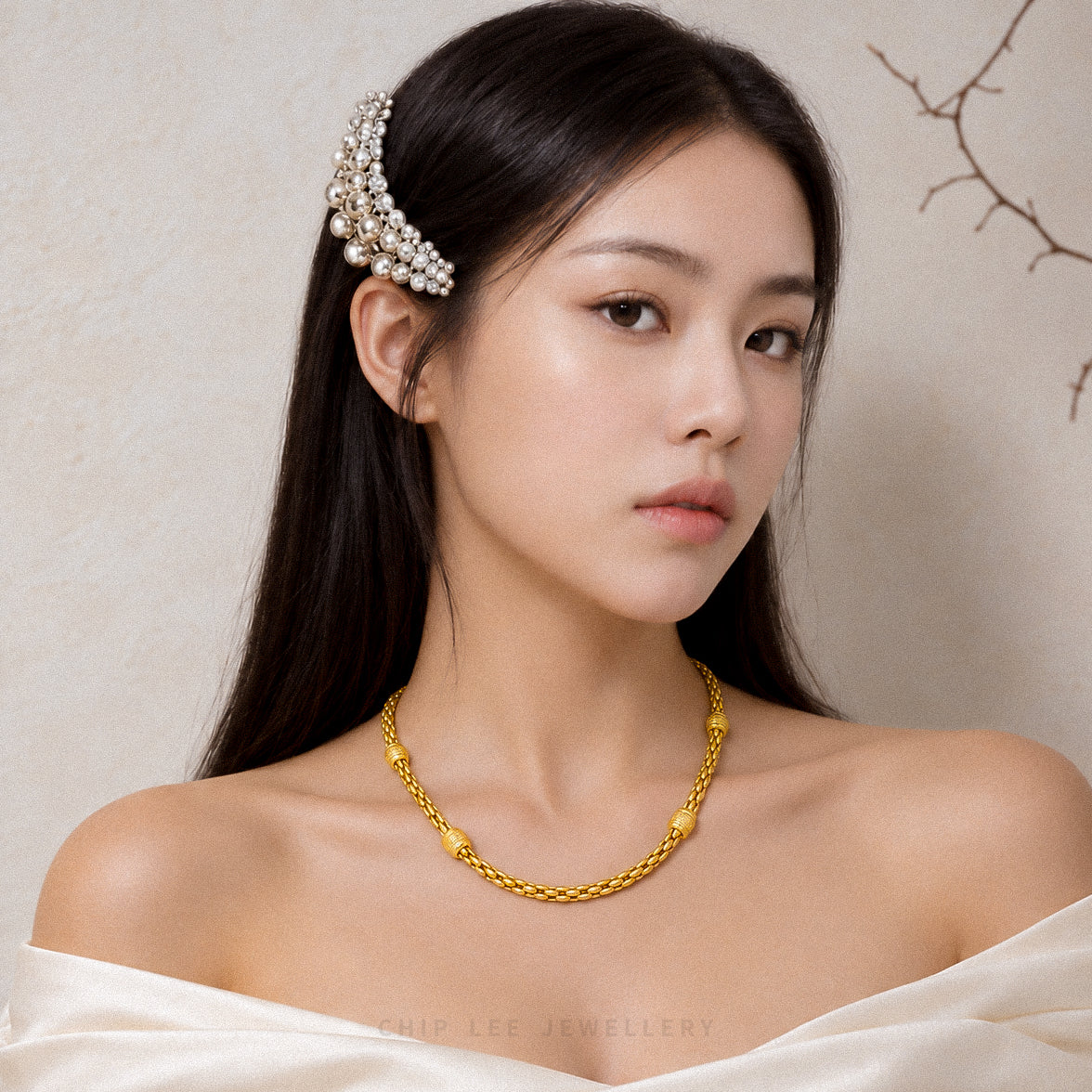 A woman with long dark hair wears a pearl and gold hair accessory and a gold chain necklace, photographed against a soft beige textured wall with a branch illustration in the corner.