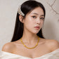 A woman with long dark hair wears a pearl and gold hair accessory and a gold chain necklace, photographed against a soft beige textured wall with a branch illustration in the corner.