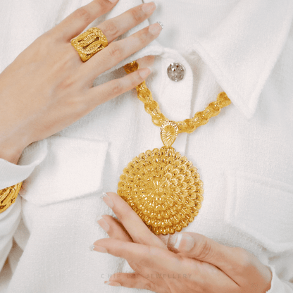 Gold necklace with intricate design held by a hand wearing a gold ring on a white background