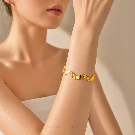 A close-up of a model's wrist wearing a gold wavy cuff bracelet with textured and polished segments, set against a soft beige background.